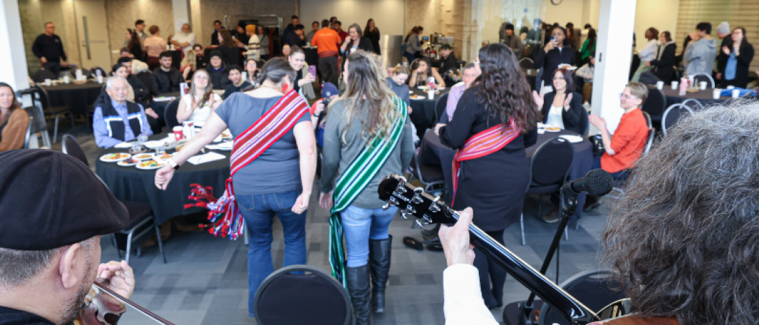 Community members gather for a Louis Riel Day celebration with live fiddle music and Métis sashes in a campus hall.