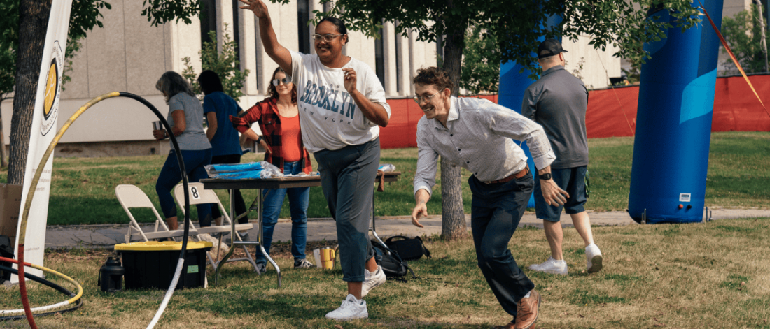 Two people play a lawn game outdoors during a campus event, tossing rings toward a target while others watch and help at a nearby table