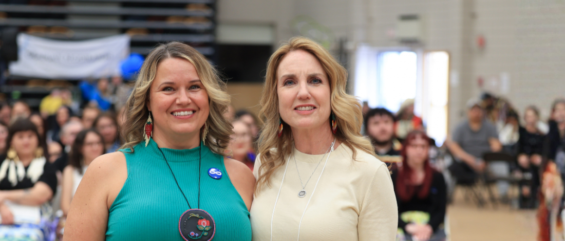 Two women standing together at a community event with an audience in the background.