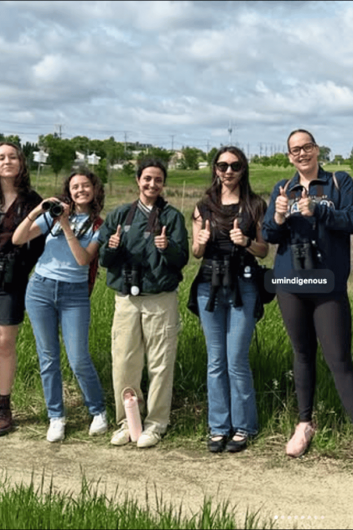 A group of students standing outdoors with binoculars, smiling and giving thumbs up during a birding activity.