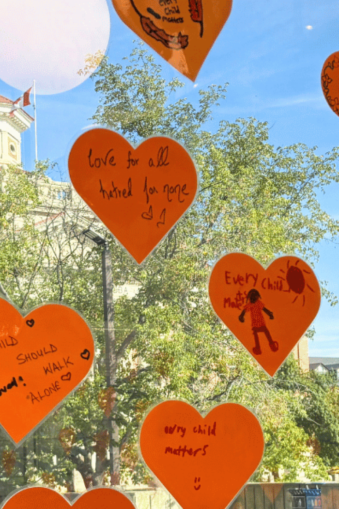 Orange heart-shaped messages on a window, including “Every Child Matters,” with the University of Manitoba campus visible outside.