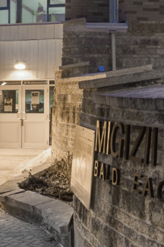A curved stone wall bearing the name “Migizii Agamik – Bald Eagle Lodge” beside a lit walkway leading to the building’s entrance at dusk.