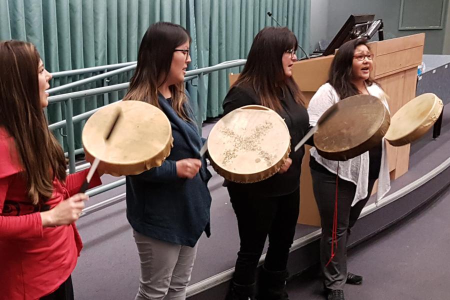 Four women stand in a row together singing and beating drums. 