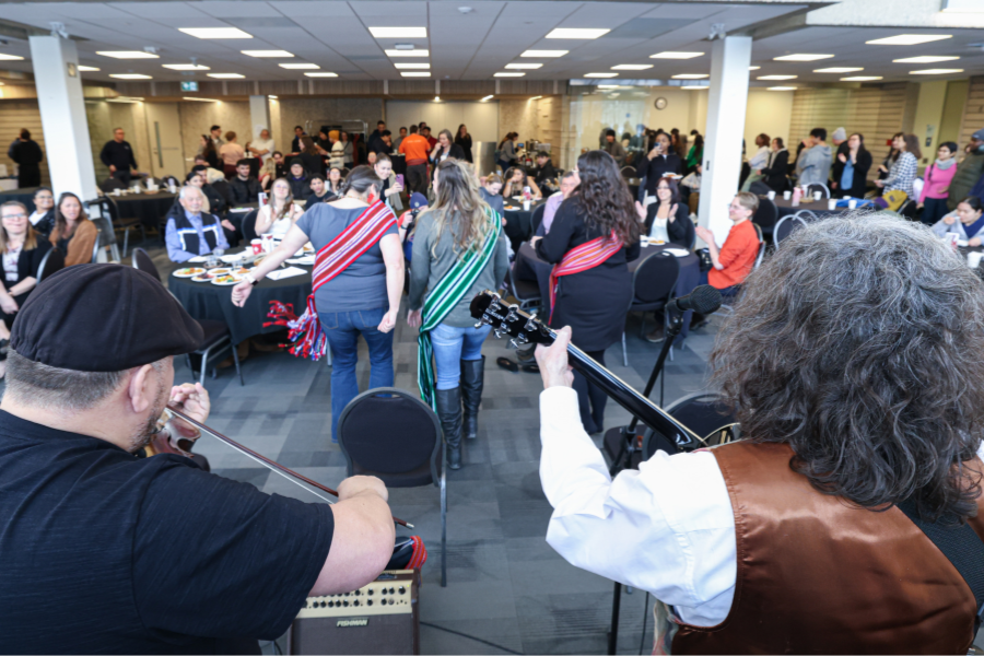 Community members gather for a Louis Riel Day celebration with live fiddle music in a campus hall.
