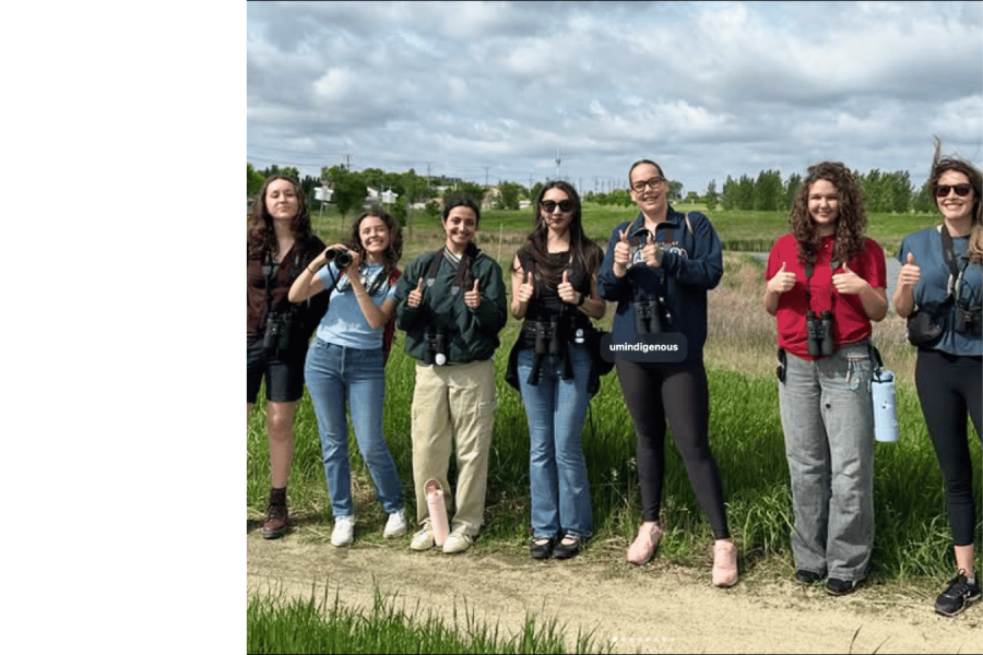 A group of students standing outdoors with binoculars, smiling and giving thumbs up during a birding activity.