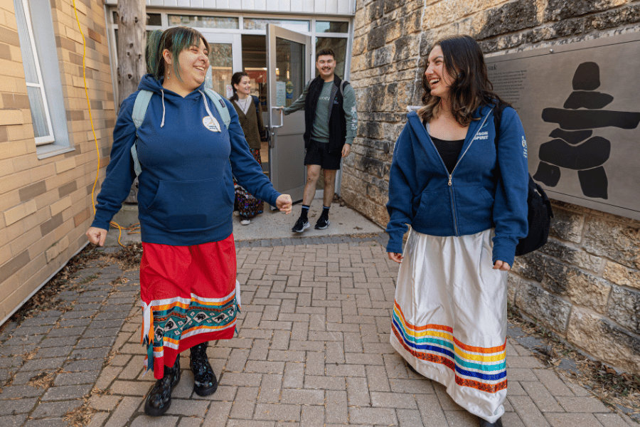 Two Bison Spirit students in ribbon skirts walk outside Migizii Agamik.