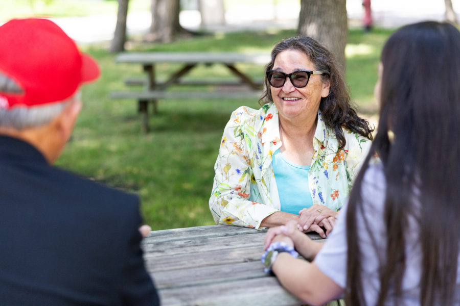 Grandmother-in-Residence sits outside at a picnic table talking to others.