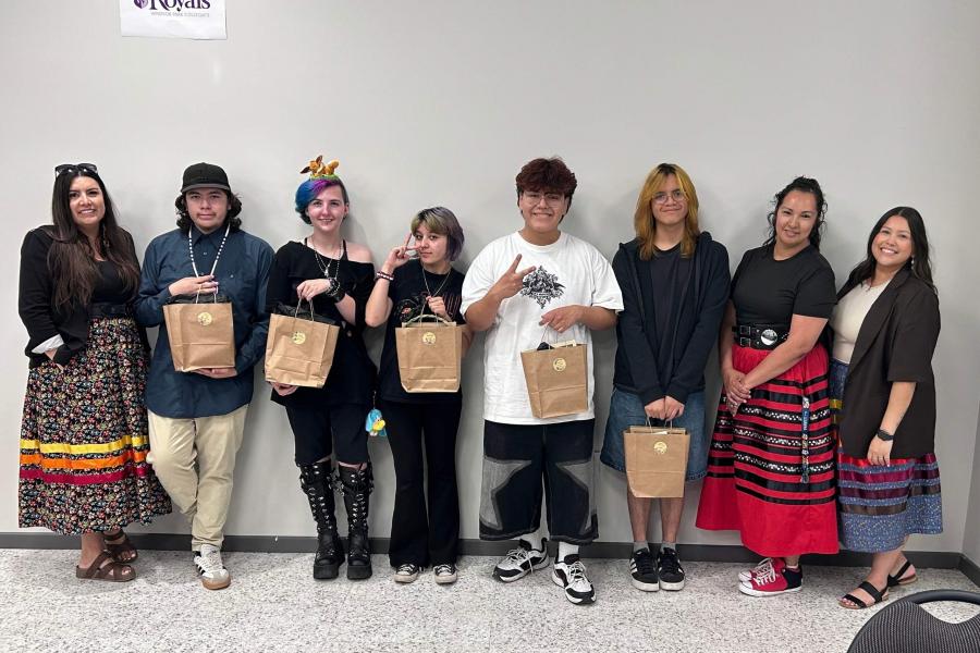 A group of people stand in front of a wall, they are posing with brown paper bags.