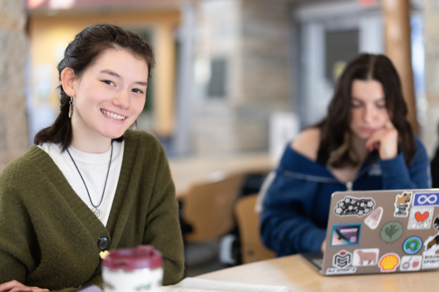A smiling student sits at a table while another works on a laptop behind her.
