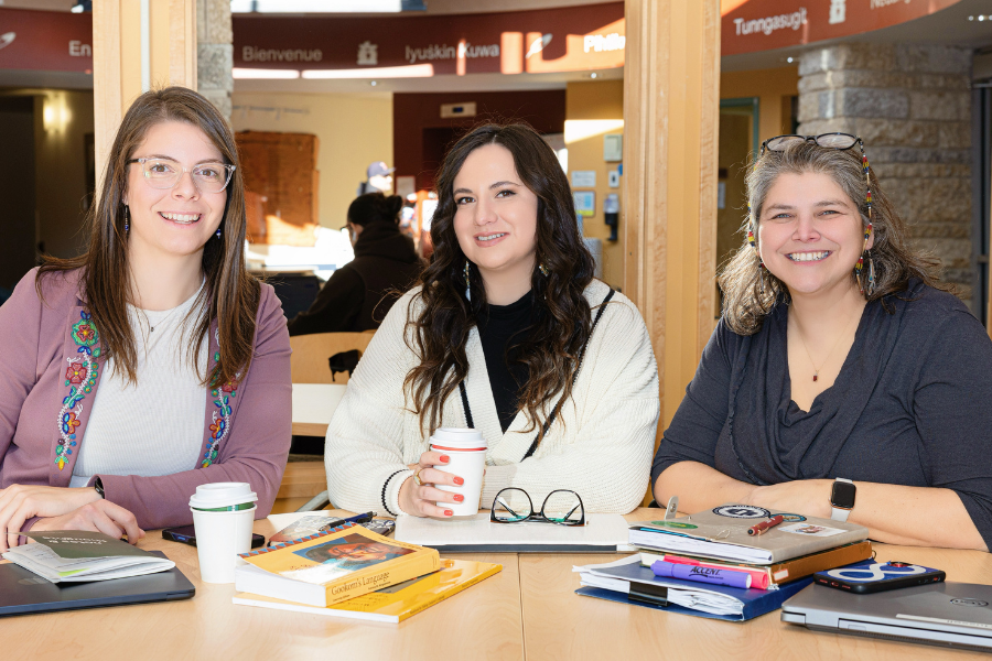Three people smile at a table with books, notebooks and coffee.