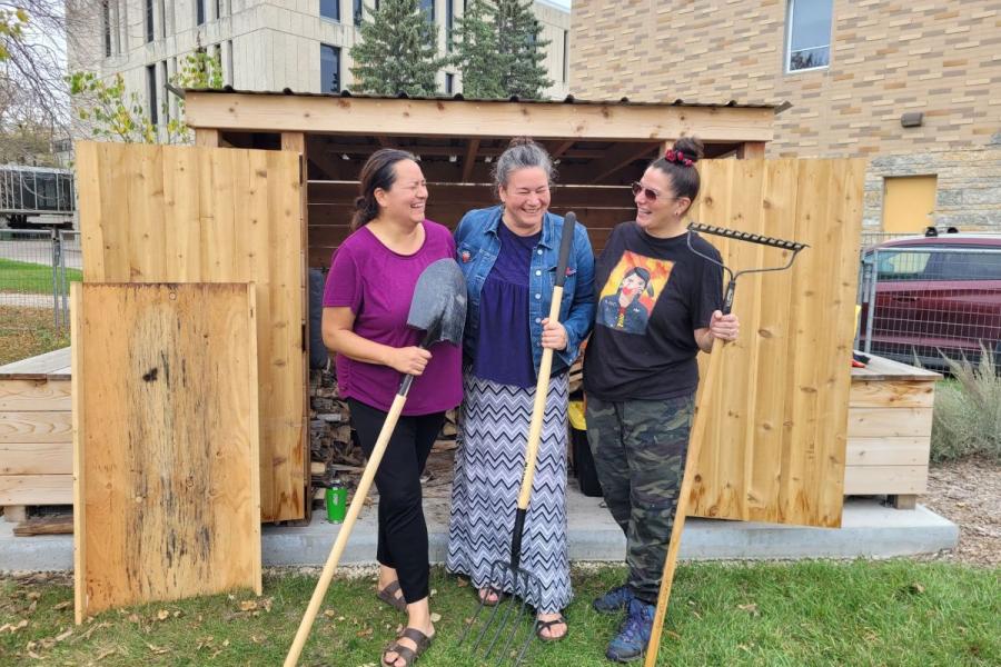 Three people stand outside, laughing together and holding large tools in front of a wooden shed.