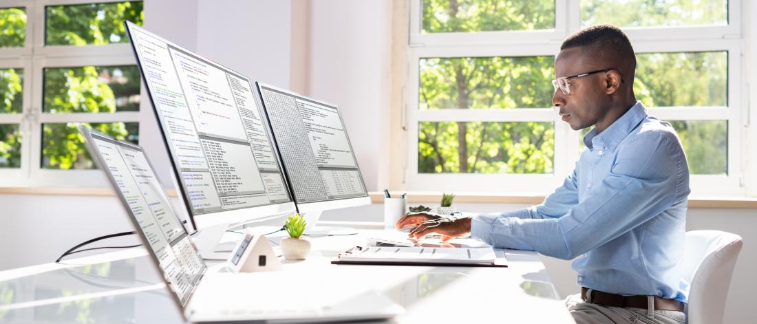 Professor working with at desk on laptop with multiple screens