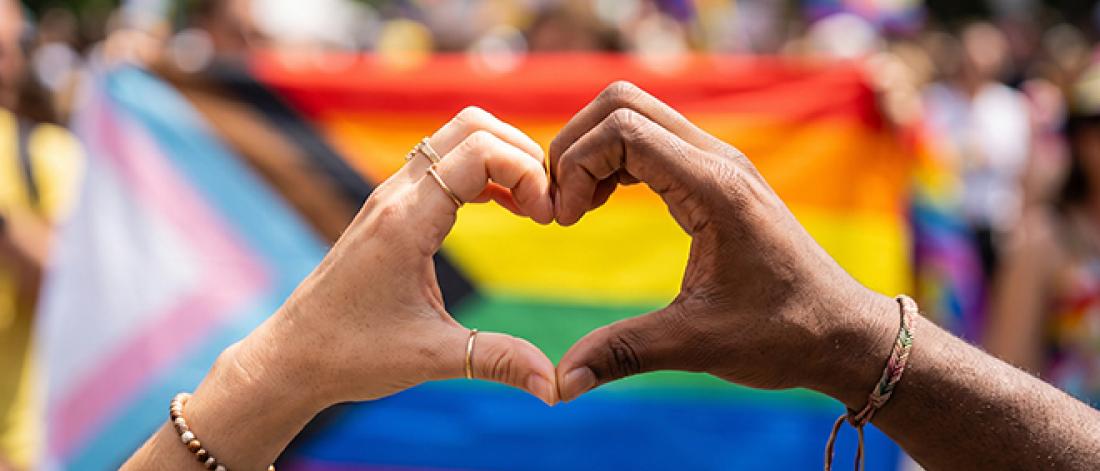 Two hands make a heart shape in front of the pride flag. One hand is caucasian and the other is African American.