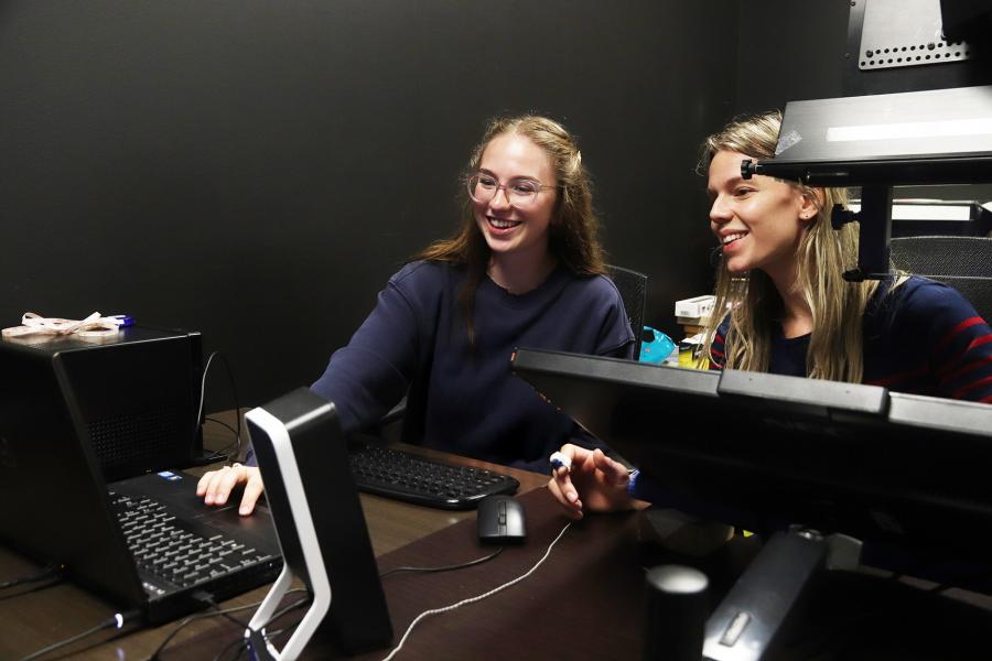 Two women sitting a desk examining information on a laptop surrounded by equipment