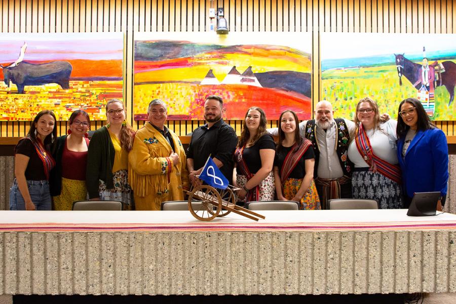 A row of people in ribbon skirts and Metis sashes stand between the Linus Wood triptych paintings and the concrete bench or Robson Hall's moot courtroom upon which a long Metis sash is laid, and a mini red river cart with Metis flag.