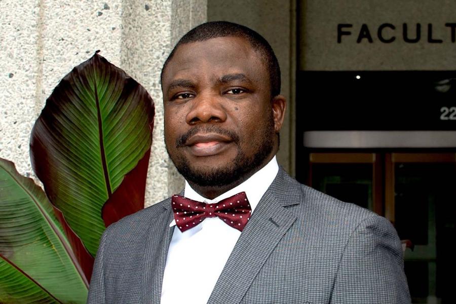a man in a grey suit with burgundy bow tie stands in front of a plant in front of the Faculty of Law building