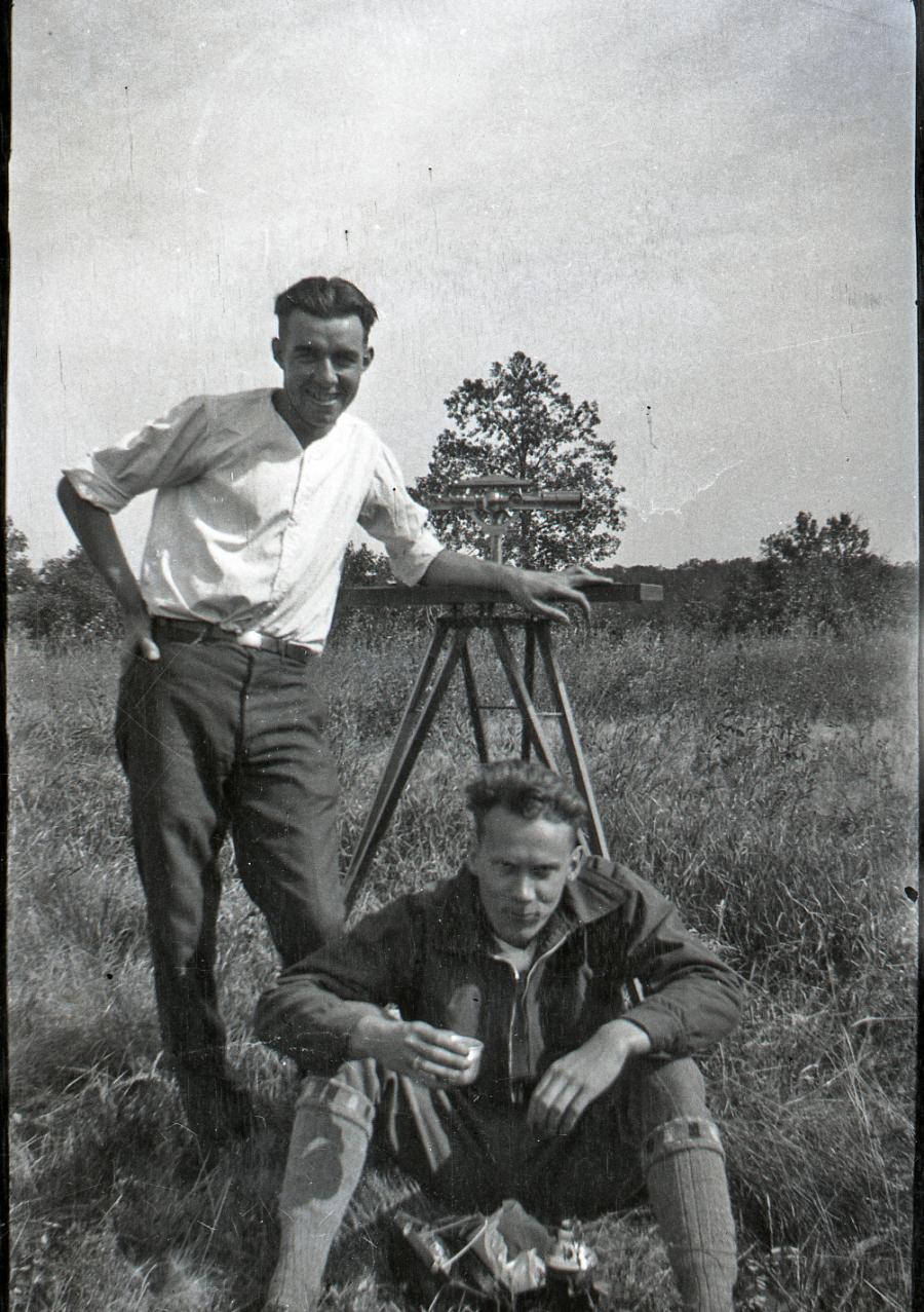 A photograph negative of two men posing with a surveying instrument while eating lunch. The man standing is Andrew Taylor, 1930. Andrew Taylor fonds, A1993-089, Box 1, File 7, Item 2