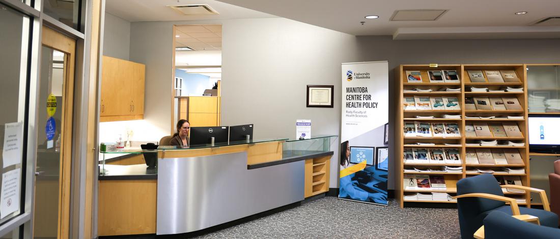 Front desk reception are at Manitoba Centre for Health Policy (MCHP). A person sits at the front desk working in front of 2 computer monitors. An MCHP banner off to the side.