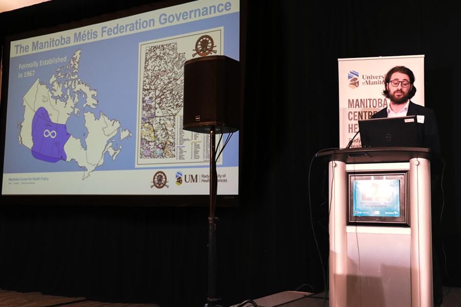 Colton Pitras at podium with presentation showing Metis territory overtop of map of Canada