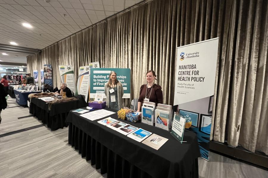 Dr. Selena Randall and Diane Gordon Pappas smiling, and working behind the MCHP booth. There are signs, and neatly organized literature lined up on the table. 