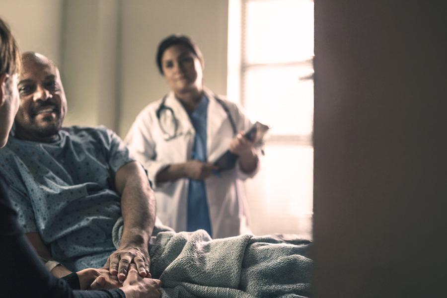 A man in a hospital bed and gown holds hands with his partner, looking at her optimistically, while a Female Physician in a white coat, wearing a stethescope, and holding a clipboard looks on from the background. 