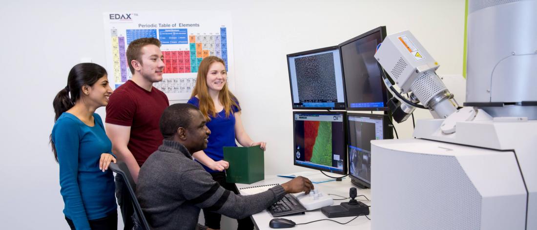 Three students gather around a fourth as he sits at a desk and operates a Manitoba Institute for Materials instrument. 