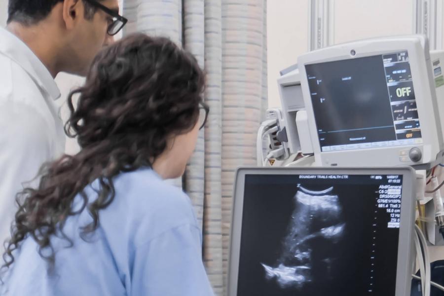 Two medical professionals look at a monitor displaying a black and white image.