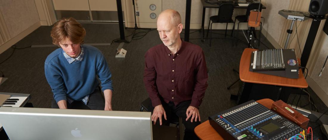 Two people seated at a desk inside the electro-acoustic composition lab surrounded by various equipment.