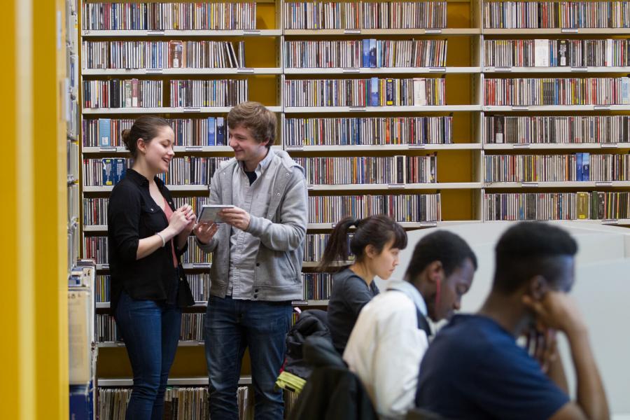 Students stand and talk in front of a large shelf of music while other students are seated and studying inside the music library. 