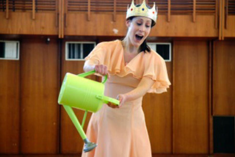 A young woman wearing a crown and carrying a watering can sings opera.