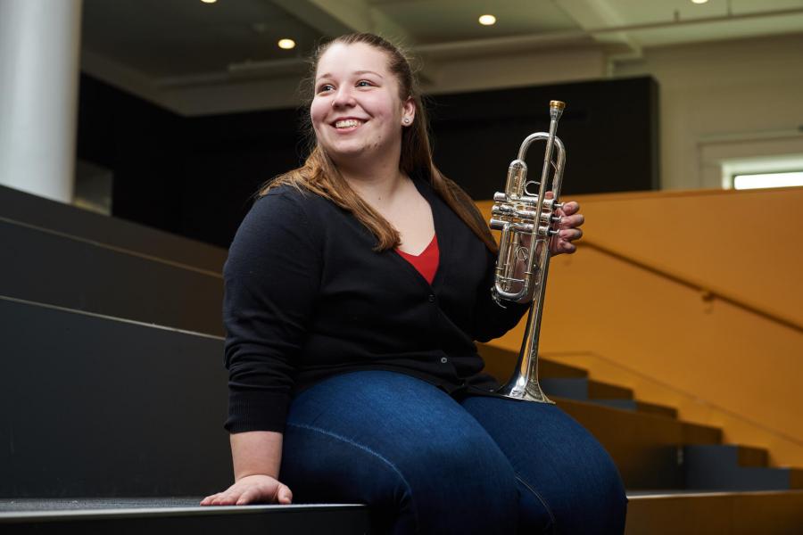 A smiling student sits in the Music library concourse holding a trumpet.