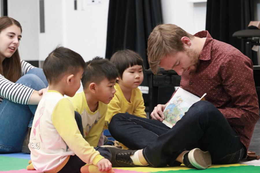 An instructor sits on the floor with small children reading a book.