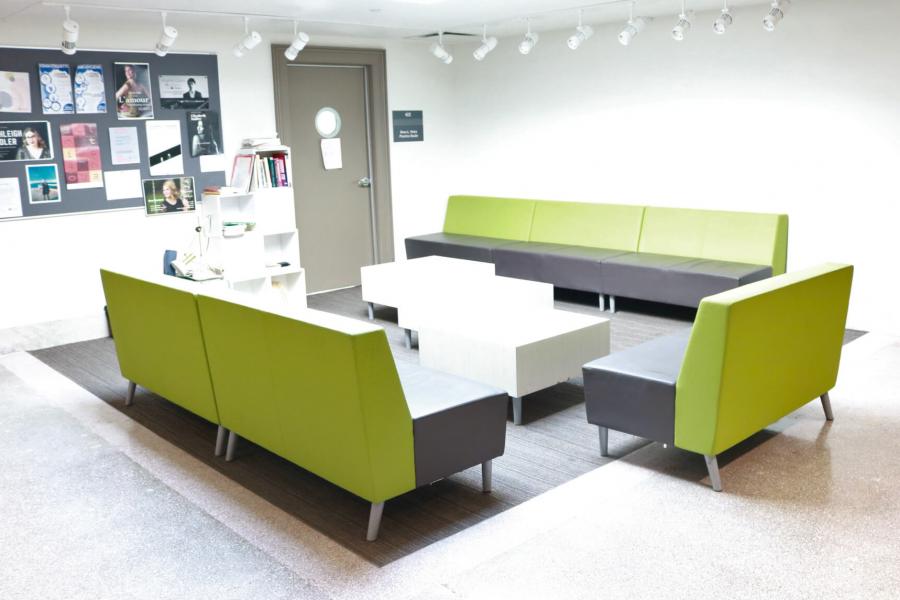 A brightly lit white room with a cork board for posters, three bright green and grey couches surrounding a carpet and three square white coffee tables.