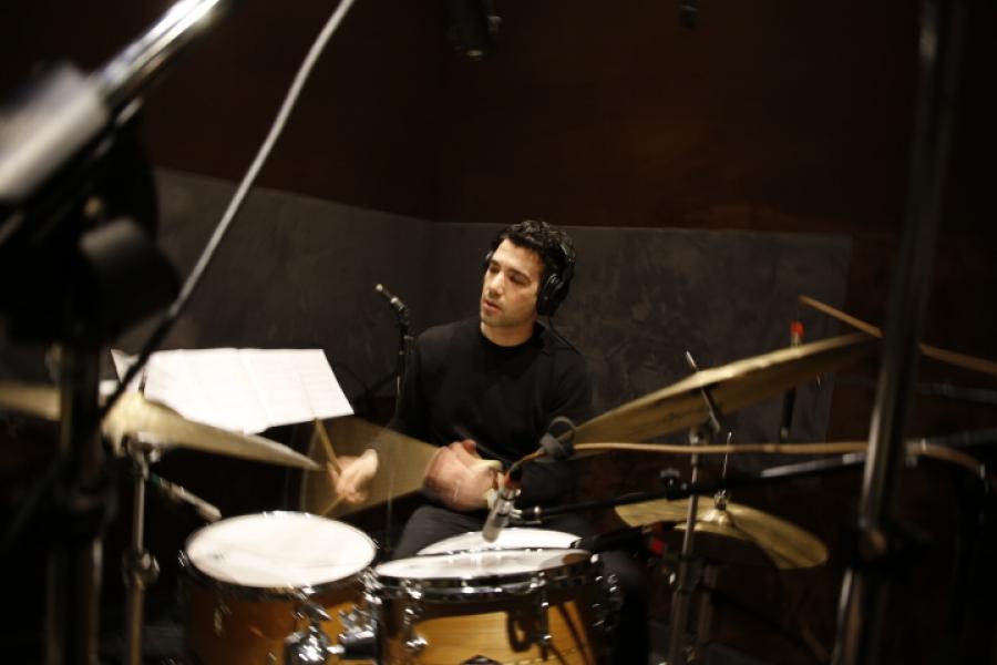 Jazz percussion professor Fabio Ragnelli sits at a drum set wearing headphones. The percussion sticks appear blurry due to the rapid music tempo Fabio is using.