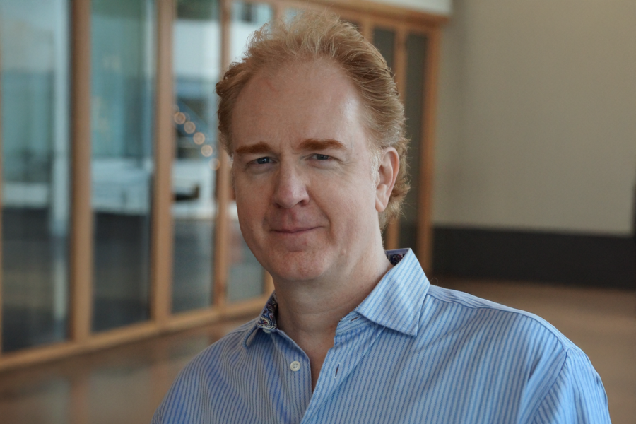 Dr. Ian Hodges, smiling, in a blue-and-white striped collared shirt, standing in a bright hallway with wood-framed glass windows behind him.
