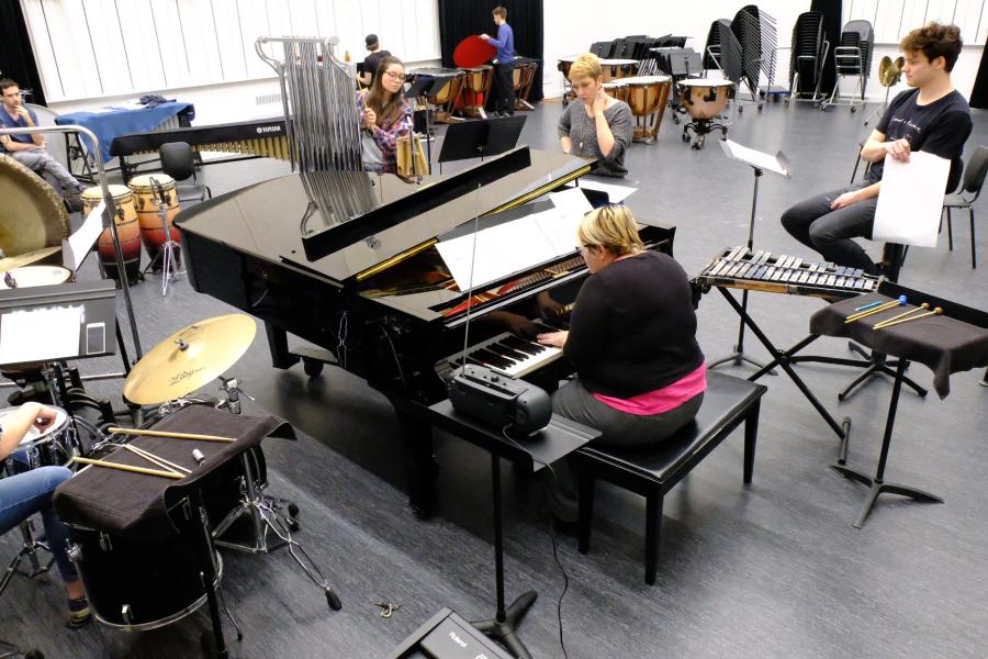 Music students rehearse with piano and percussion instruments in an ensemble classroom.