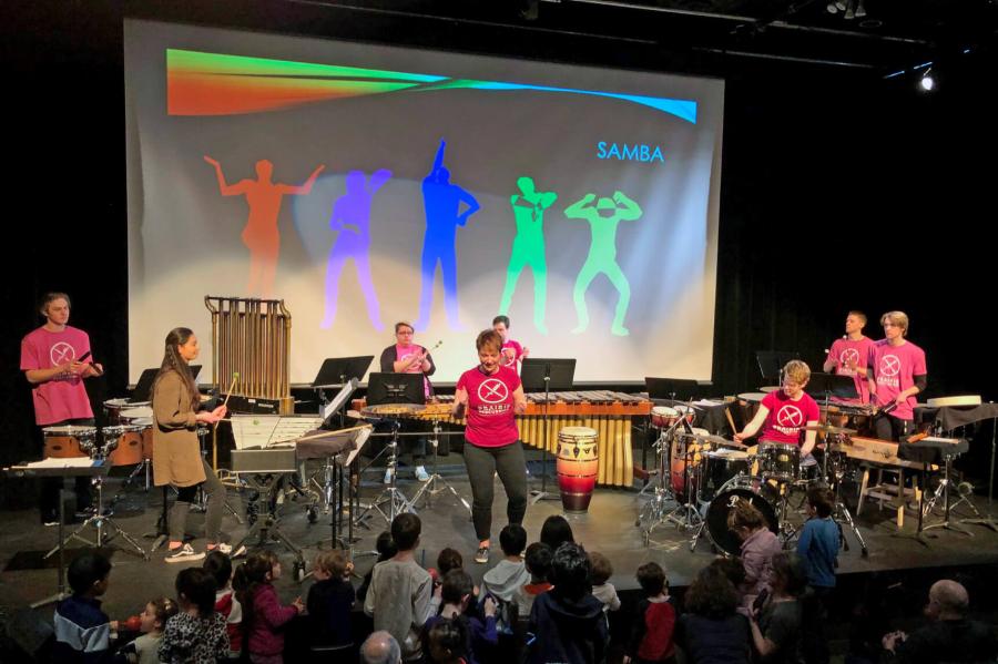 A crowd of people watch a performance on a stage with eight performers playing various musical instruments and a large screen behind them with colourful graphics projected onto it. 