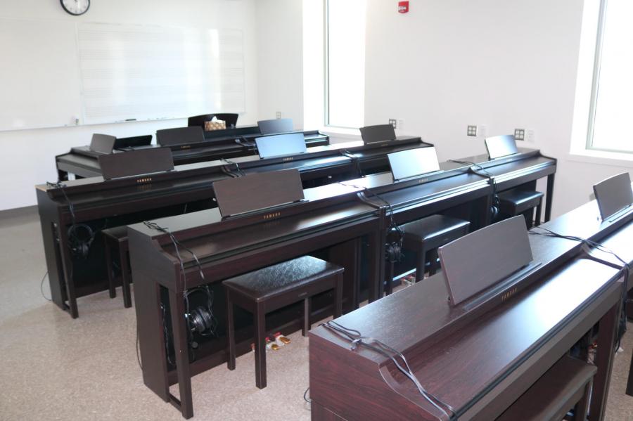 A room with a wall lined with tall narrow windows and another with a large white board. Multiple rows of three Yamaha keyboards fill the space.