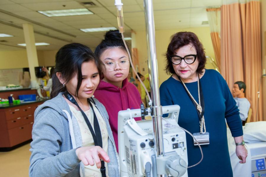 Two nursing students look at a monitor, an instructor standing nearby points at the monitor screen.