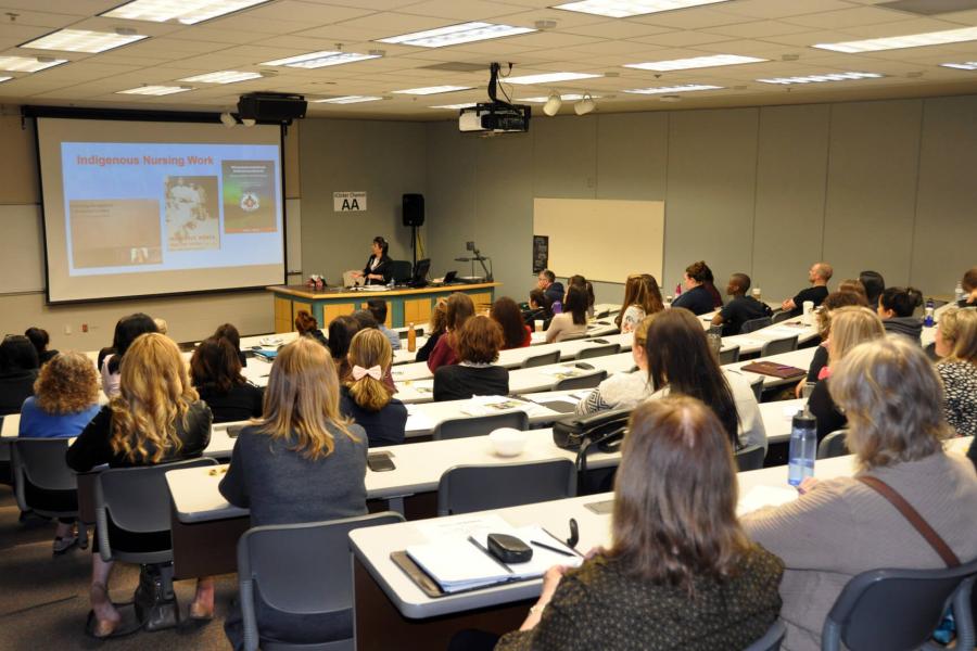 A classroom of Master of Nursing program students watch a presentation on a projector screen.