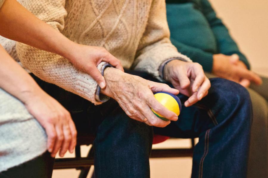 A woman gently holds the wrist of an elderly patient who is seated and holding a ball.