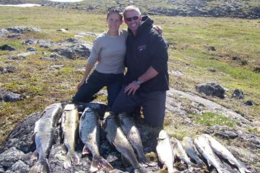 Two people standing on a riverbank with freshly caught fish on the ground in front of them. 