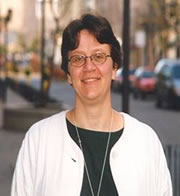 This is a picture of Deborah Stienstra. She is standing out of doors, wearing a dark green shirt and white cardigan. She is smiling, has short brown hair, brown eyes and is wearing glasses.