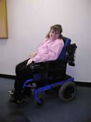 this is a picture of Heidi Janz. She is sitting in a white room with a dark floor, wearing black pants and a pink sweater. She is smiling, has long light brown hair and is sitting on an angle in a wheel chair
