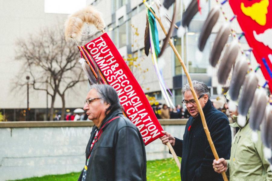 A man, Carl Stone, leading a ceremonial procession. The man beside him holds a banner that reads: Truth and Reconciliation Commission of Canada.