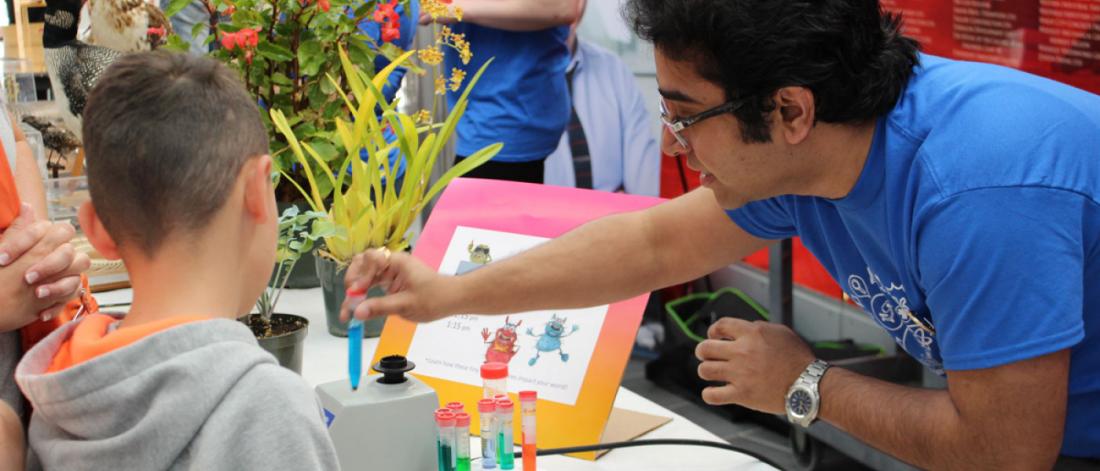 a science volunteer wearing eyeglasses and a blue shirt showing a child a test tube with blue liquid in it.