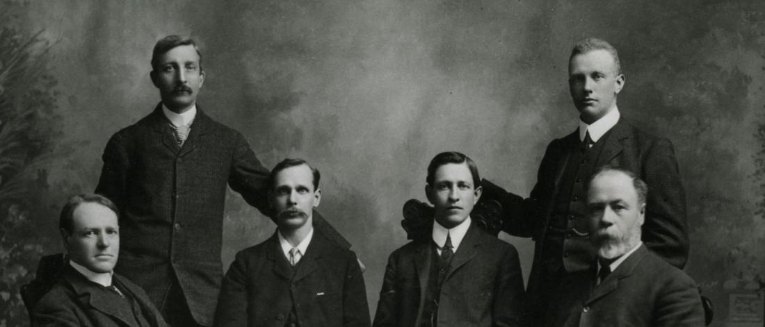 Six people two of them standing and the rest are sitting down all wearing a coat and tie.