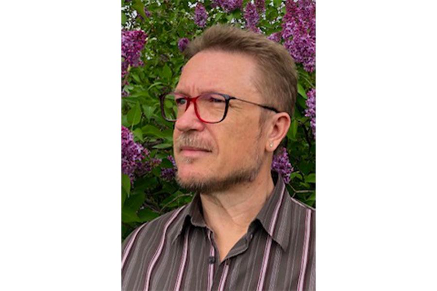 Vladimir Yurkov wearing an eyeglass looking on the left side in front of a tree with purple flowers.