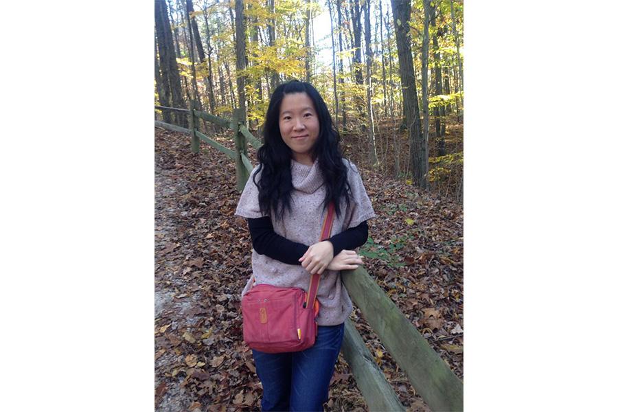 Kang Ling Liao wearing a purple sweater with a pink bag smiling at the camera in front of trees.