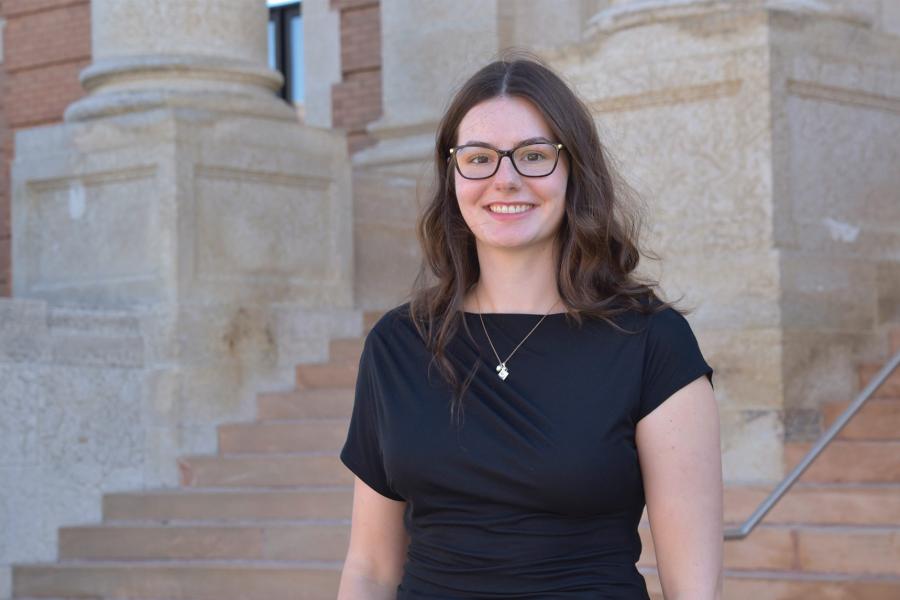 Neve Loewen wearing a black top with a pendant and eyeglass smiling at the camera.